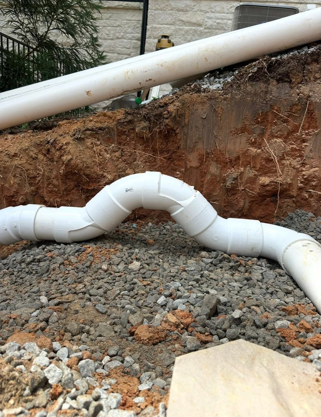 White sewer pipe bent over gravel beside an eroded dirt embankment and larger pipe above.