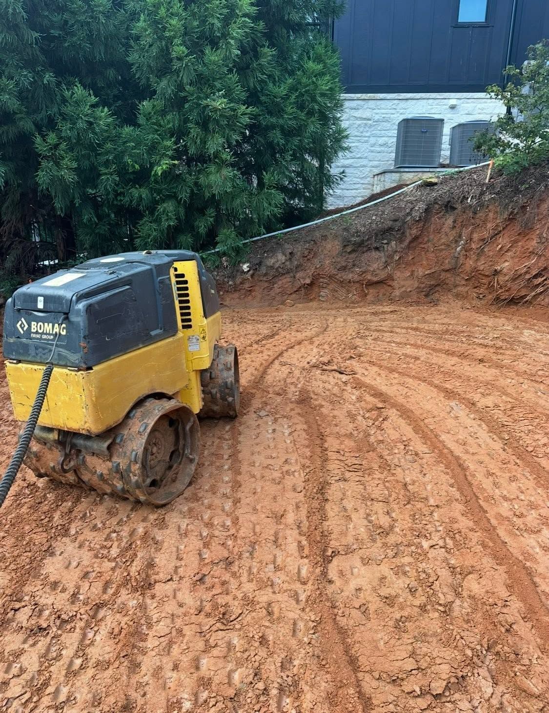 Yellow compact roller on muddy red dirt near a house and trees