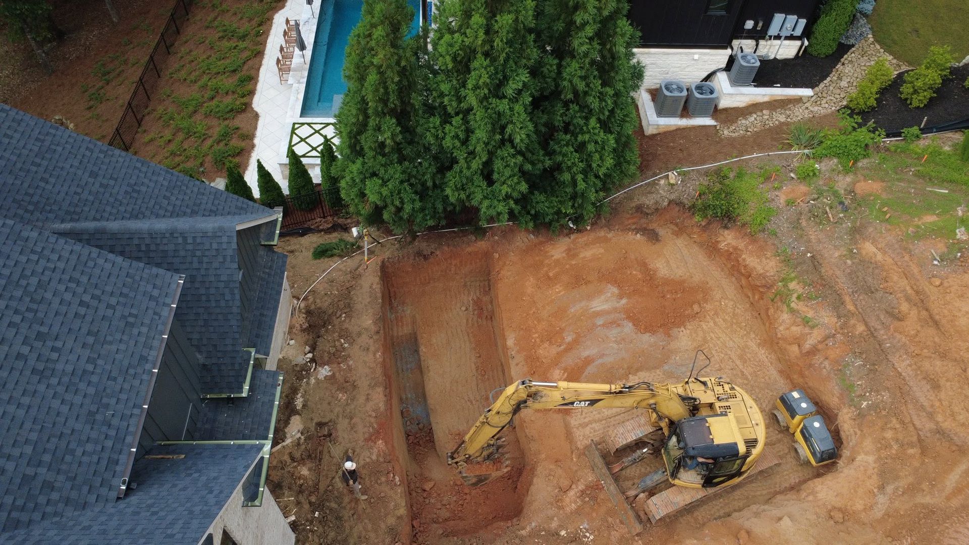 Aerial view of a yellow excavator clearing dirt beside a house and trees in a yard