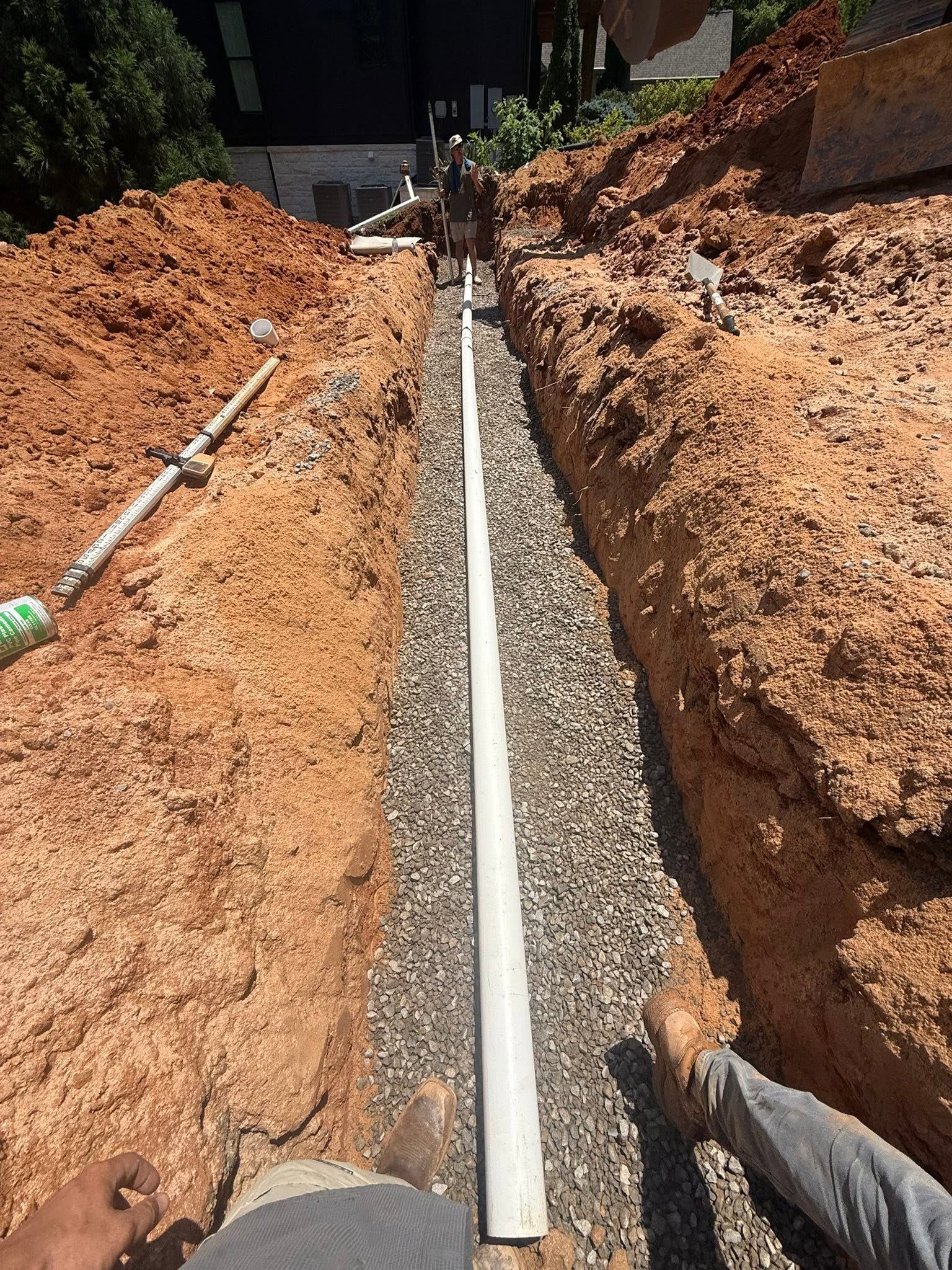 Workers installing a white drainage pipe in a deep dirt trench beside a building