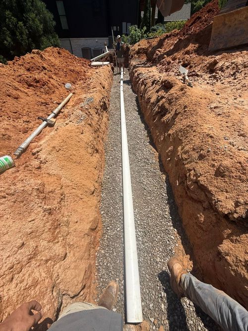 Workers installing a white drainage pipe in a deep dirt trench beside a building