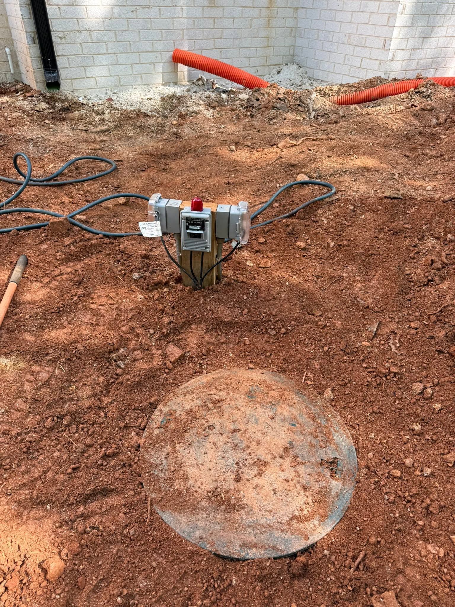 Construction trench with a utility box, exposed cables, and a round metal cover in muddy dirt near a wall
