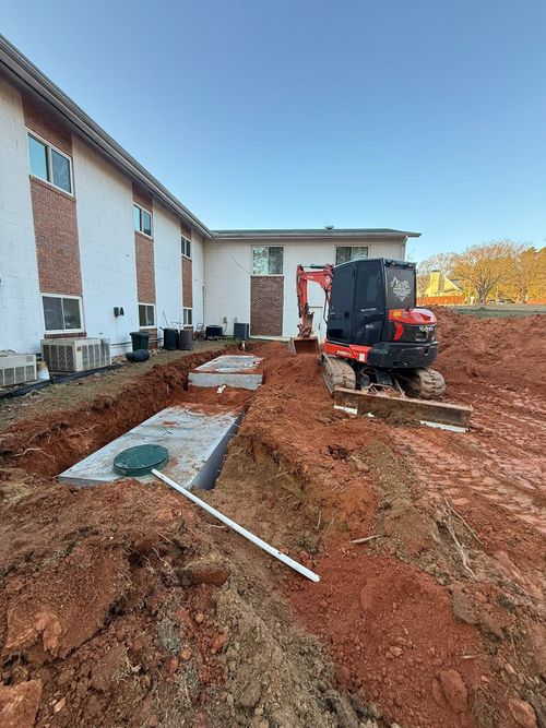Construction equipment digging muddy trench beside a building, with red soil and exposed utility lines.
