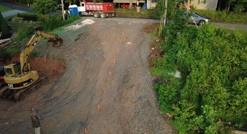 Gravel road under construction beside green vegetation, with a yellow excavator on the left.