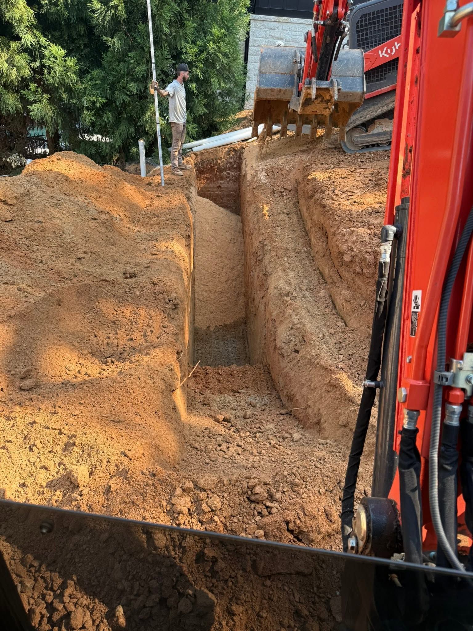 Excavator digging a trench beside a dirt road, with piles of soil and trees in the background