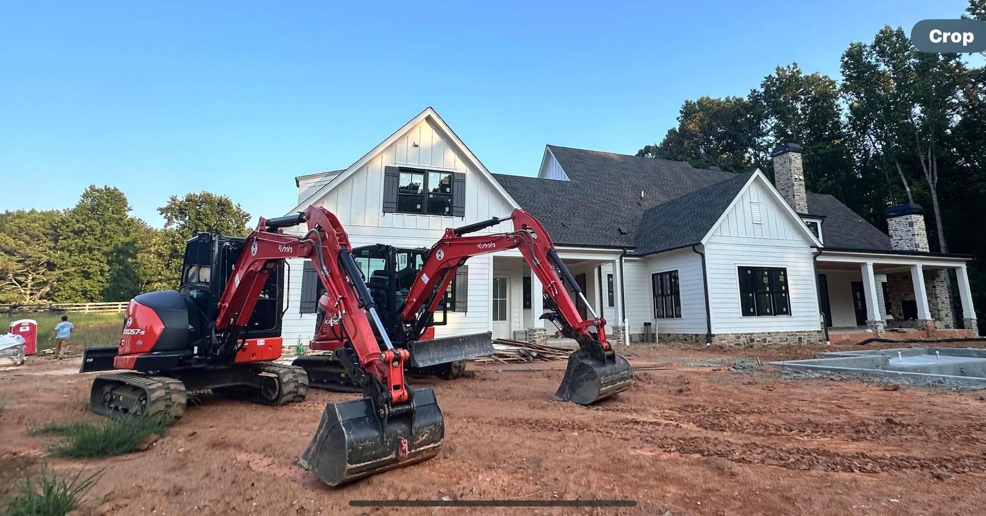 Red excavator working on dirt in front of a white house under construction.