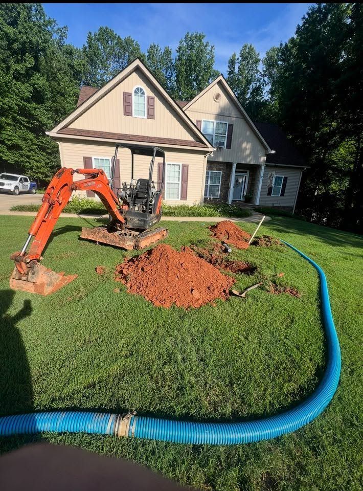 Orange excavator digging a lawn trench beside a house, with a blue hose and dirt pile in front