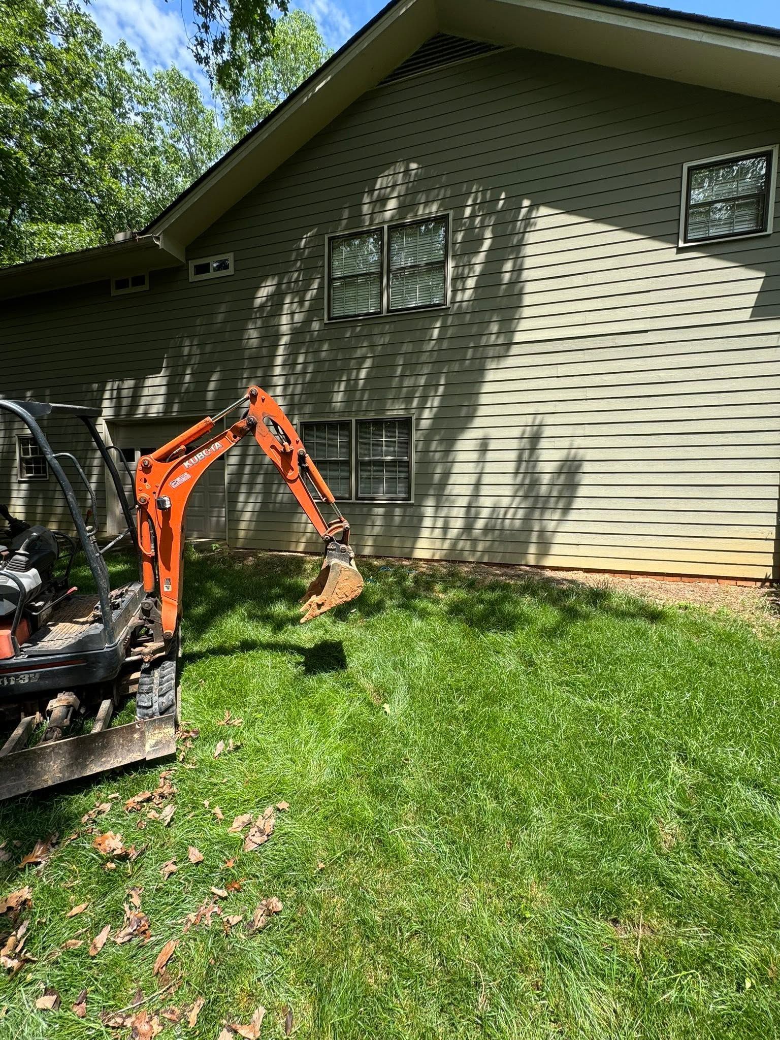 Excavator by a house’s vinyl siding, clearing grass and debris in a sunny yard.