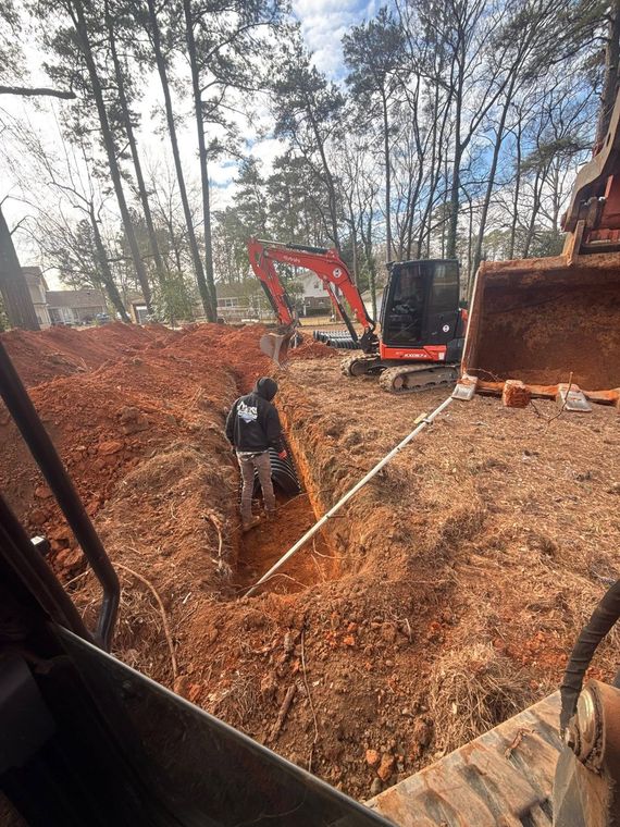 Excavator loading brown mulch in a wooded area with a worker standing nearby