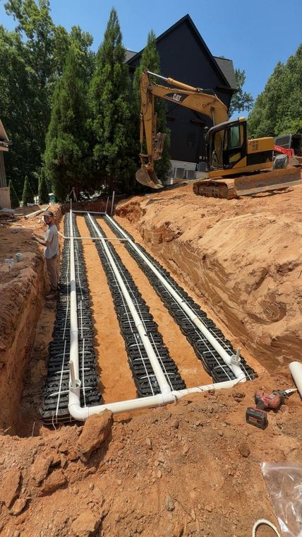 Trenched utility lines and white conduit laid in red dirt beside a house under construction.