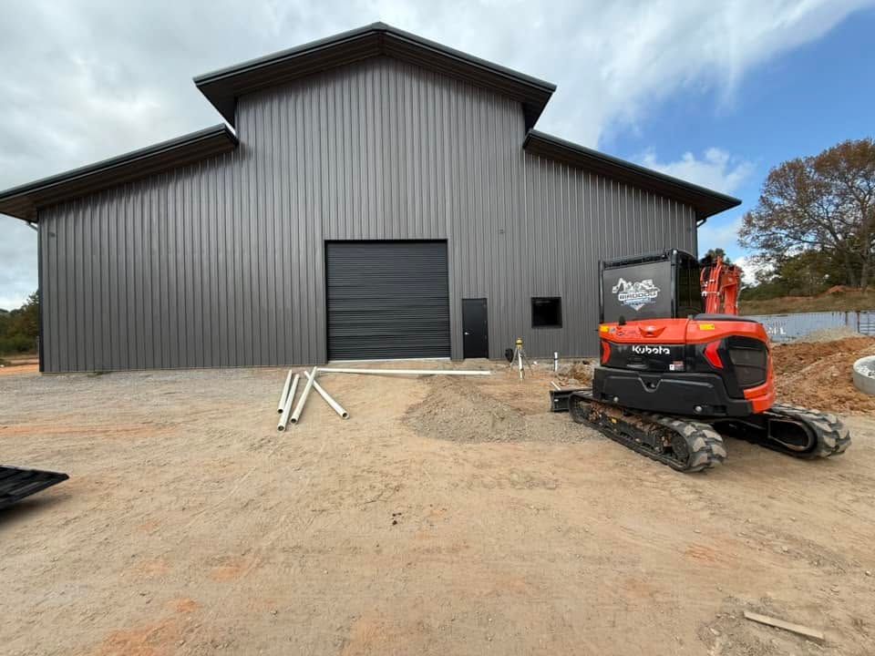 Large dark-gray barn under construction with a red tracked skid steer on a dirt lot.