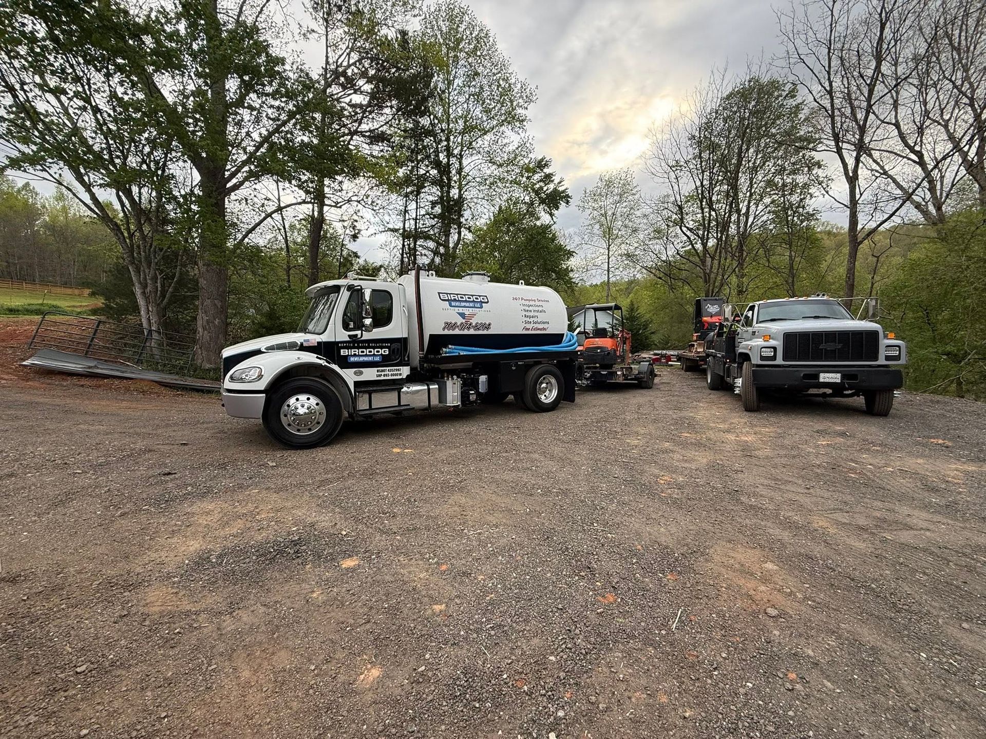 Two utility trucks parked on a gravel lot beside trees, with equipment and workers in the background