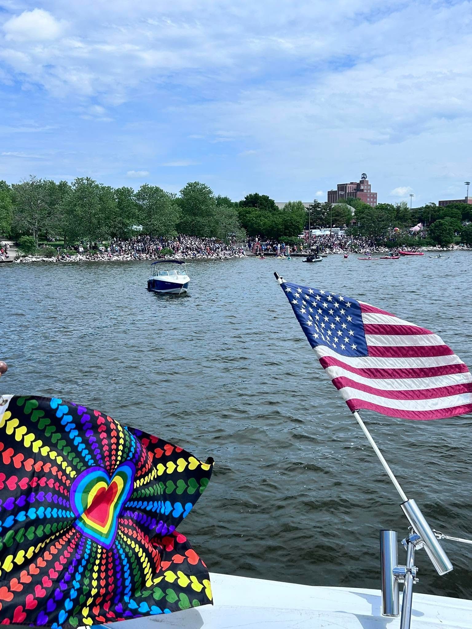 Colorful umbrella and U.S. flag on a boat, overlooking a lake with people along the shore and a cloudy sky.