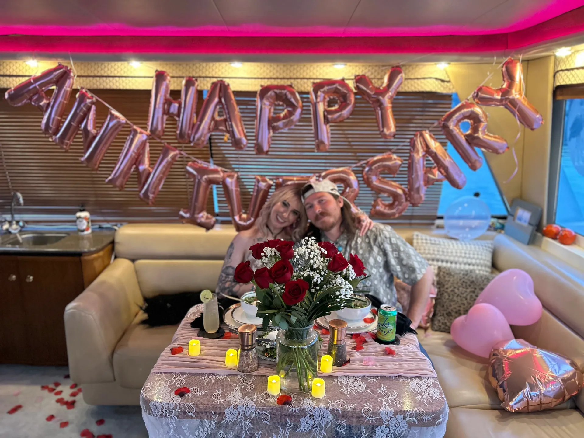 Couple seated at a decorated table with rose balloons and “Happy Anniversary” balloons in a cozy room