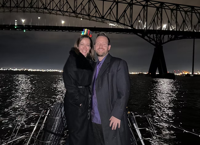 Couple smiling on a boat at night beneath a lit bridge, city lights reflecting on the water.
