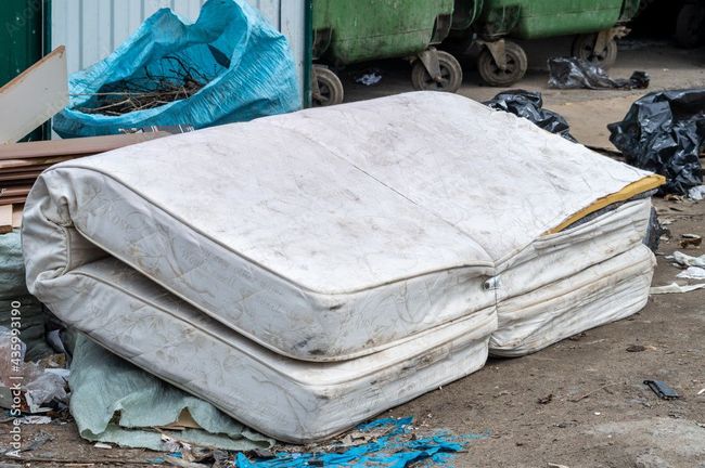 Discarded mattress on a trash pile with blue bags and green bins in the background.