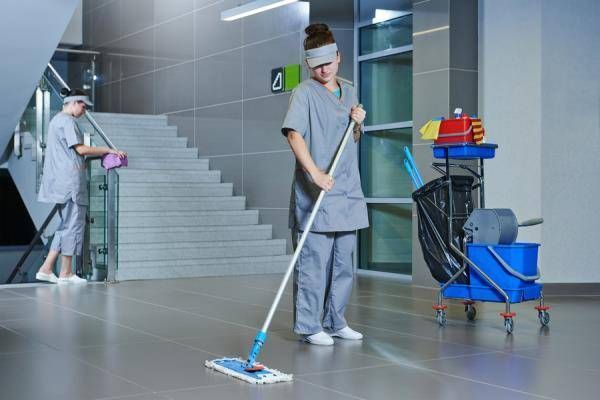 Due custodi puliscono un corridoio. Uno lava i pavimenti, l'altro spolvera le scale. Un carrello per le pulizie nelle vicinanze.