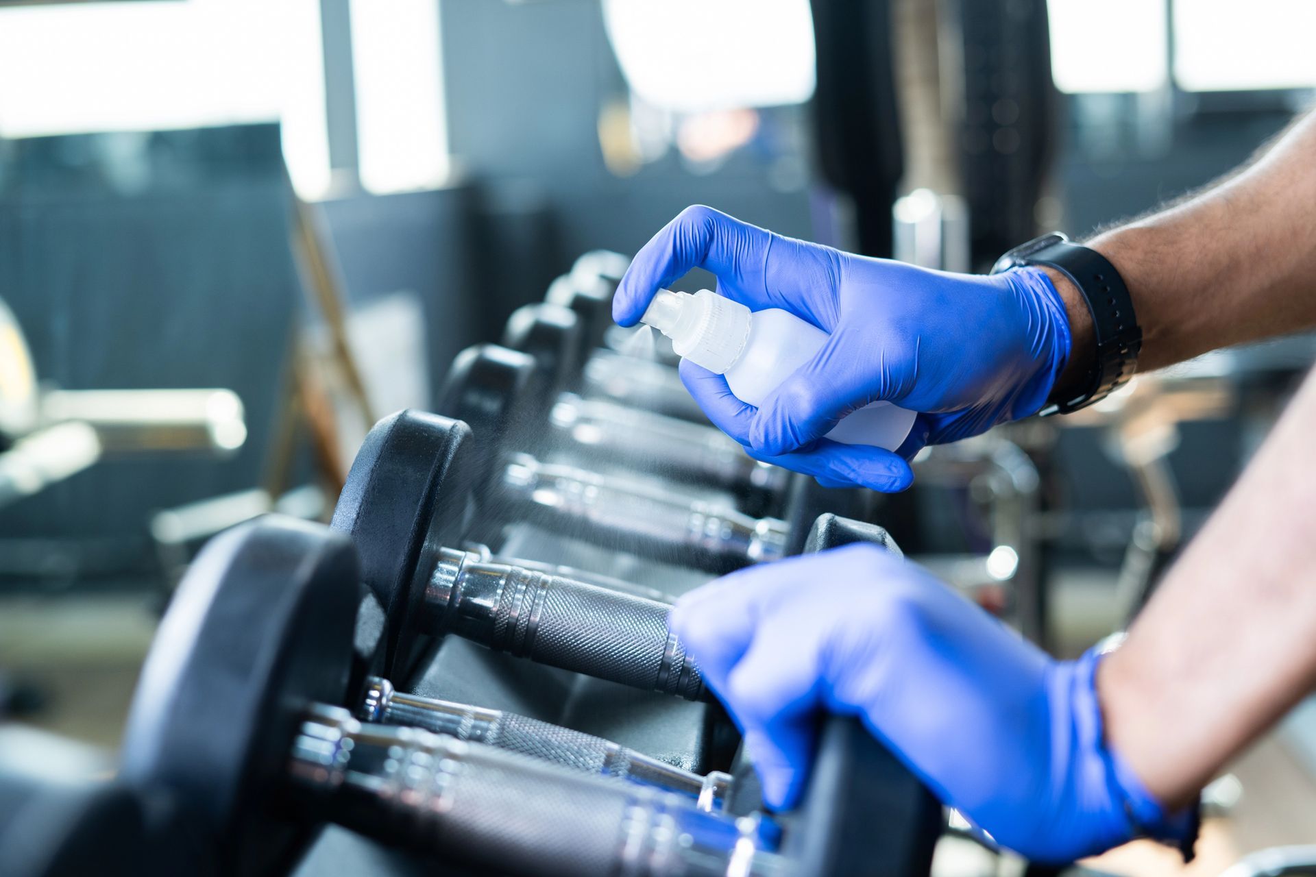 Una persona che indossa guanti blu spruzza disinfettante sui manubri in una palestra.