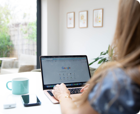 Two people working at a wooden table with laptop, notebook, and coffee. One typing, the other looking on.