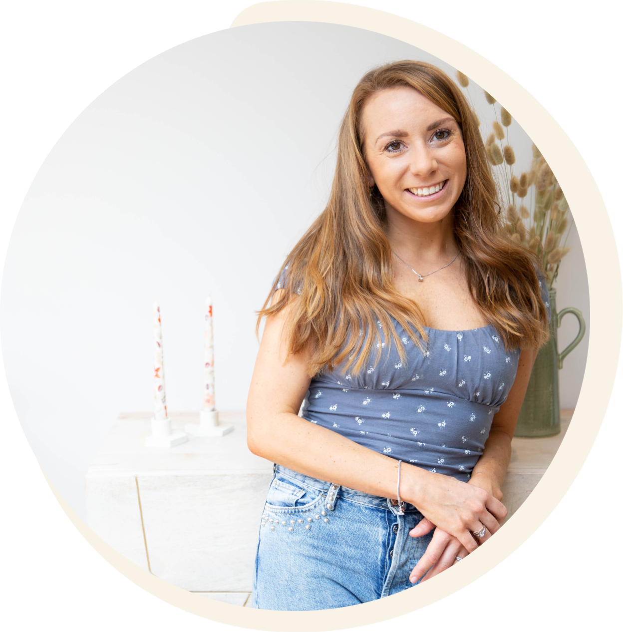 Woman smiling, leaning on a white surface with candles and dried flowers. She wears a blue top and denim shorts.