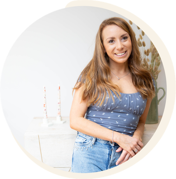 Woman smiling, leaning on a white surface with candles and dried flowers. She wears a blue top and denim shorts.