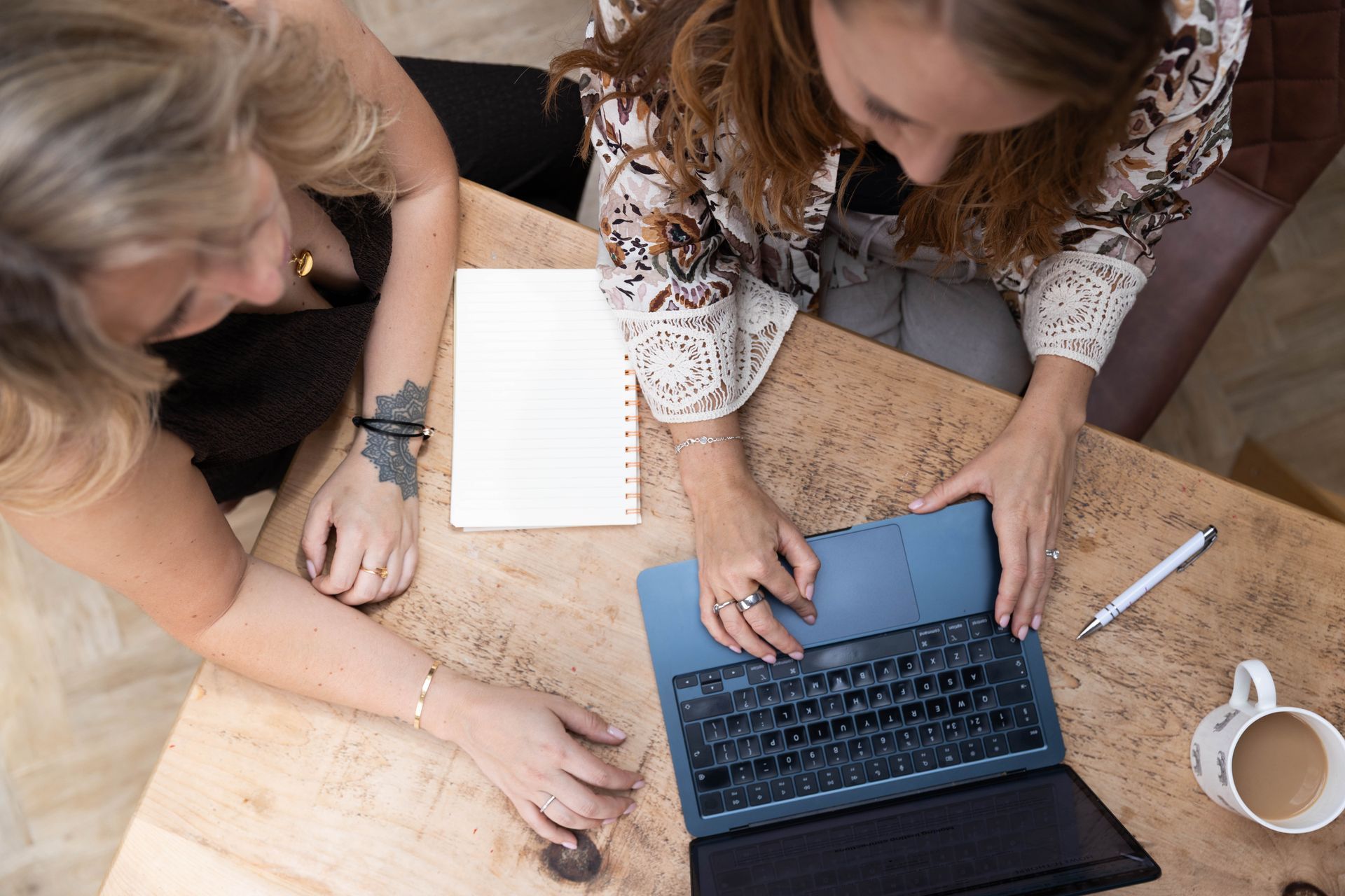Two people looking at laptop on wooden table, notebook, coffee cup, pen present.