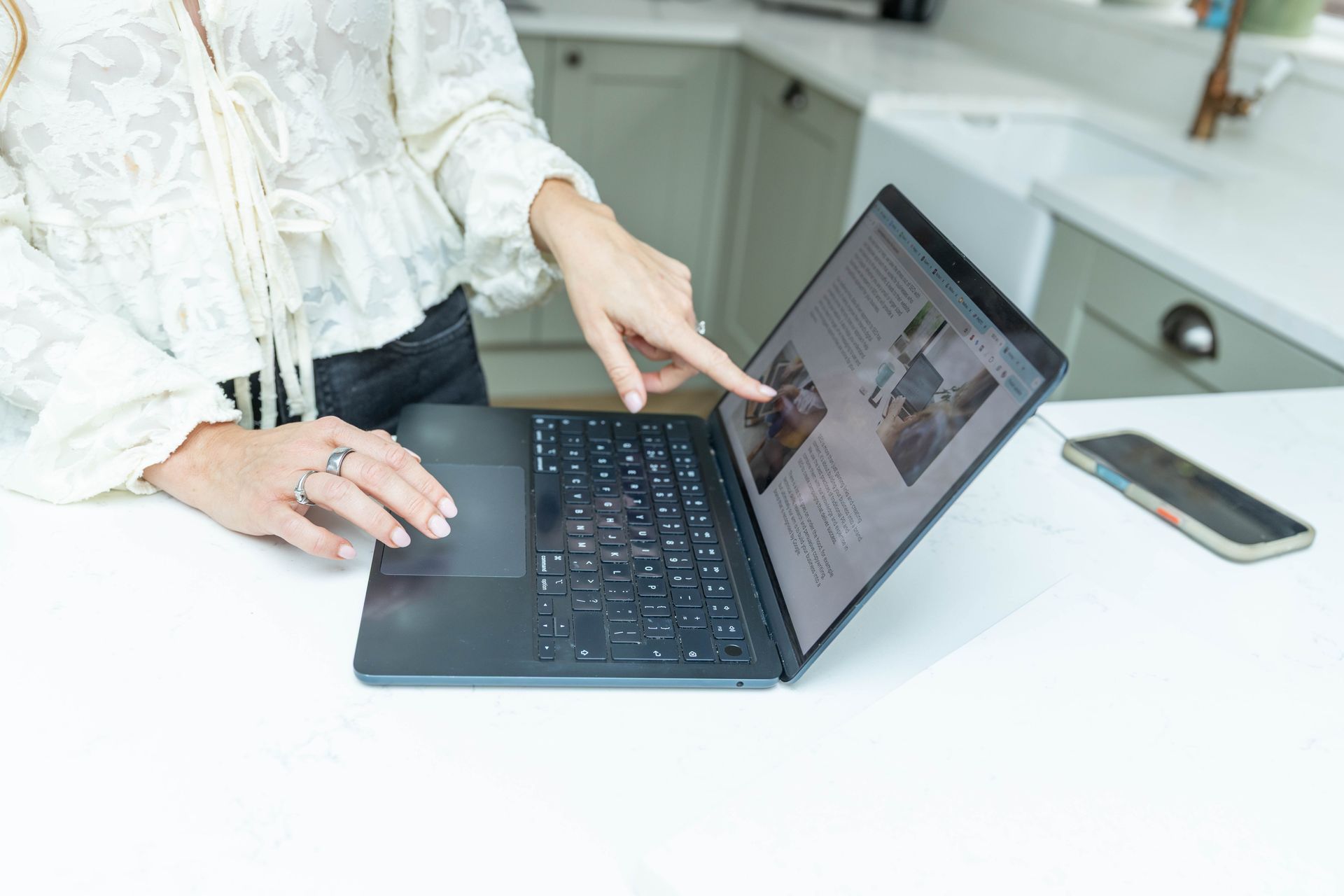 Woman points at laptop screen in a kitchen, indicating content.