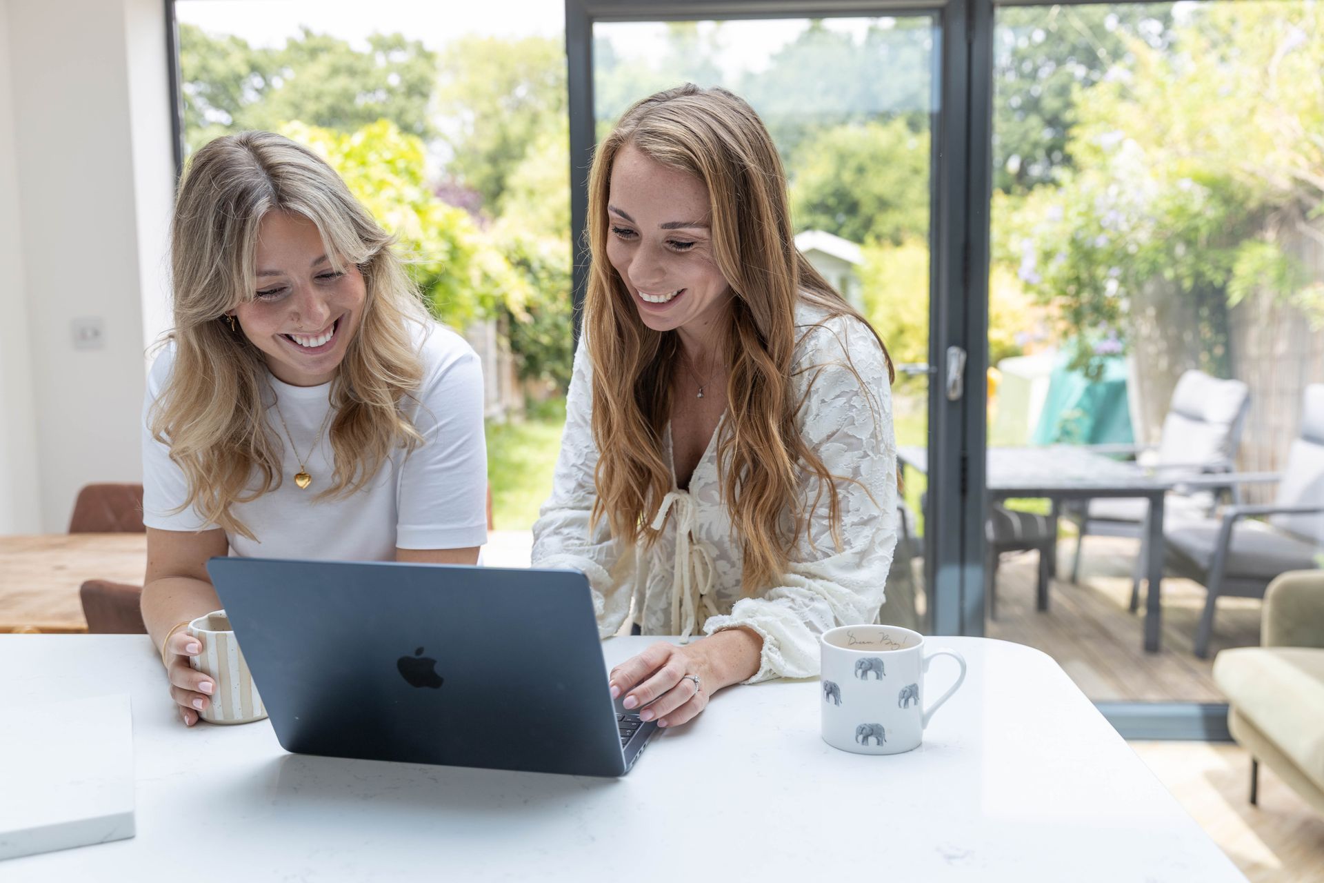 Two people collaborate at a wooden table, one using a laptop, the other with a notepad. Coffee cup present.