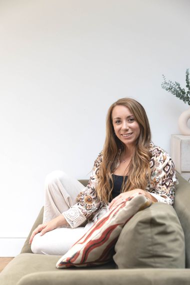 Woman smiles, sitting on a green couch, wearing patterned jacket and white pants; pillow behind her.
