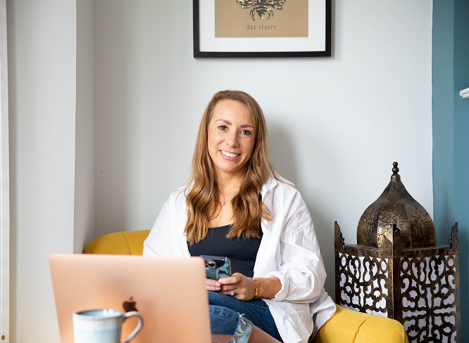 Woman in white shirt smiles, holding phone, laptop open on yellow couch. Decorative lamp beside her.