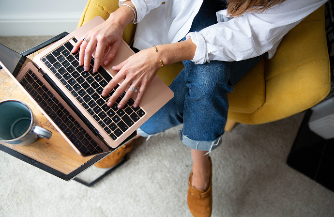 Person typing on a laptop, sitting in a yellow chair with a coffee table holding a mug.