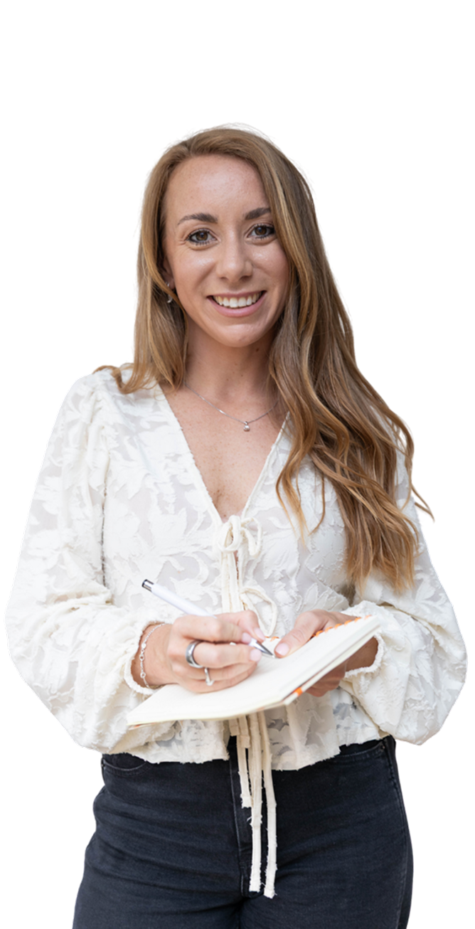 Woman smiles, holds pen and notebook. Wearing white floral blouse, black pants.