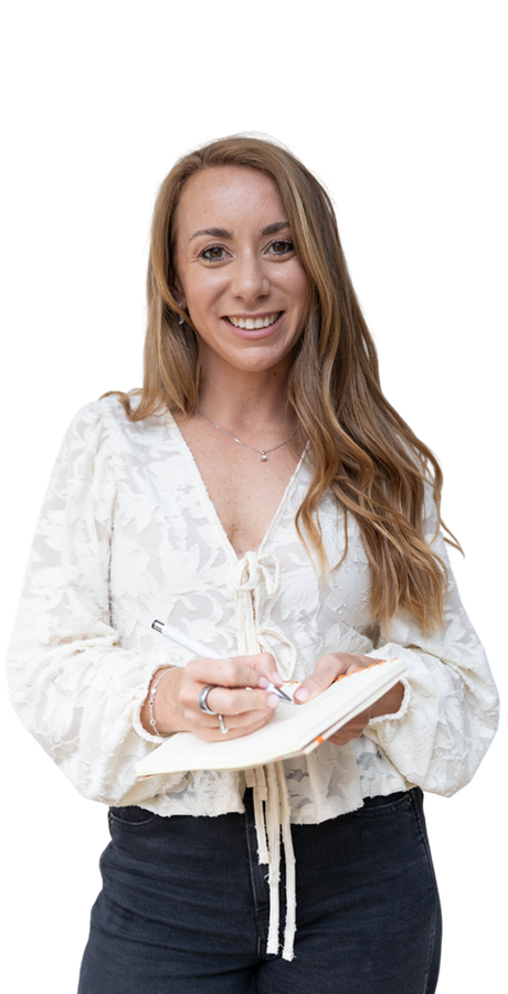 Woman with long hair, holding pen and notepad, smiling at the camera. Wearing white floral blouse and black jeans.