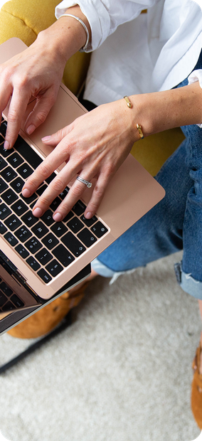 Person using a rose gold laptop while sitting on a light-colored rug, wearing jeans and a white shirt.
