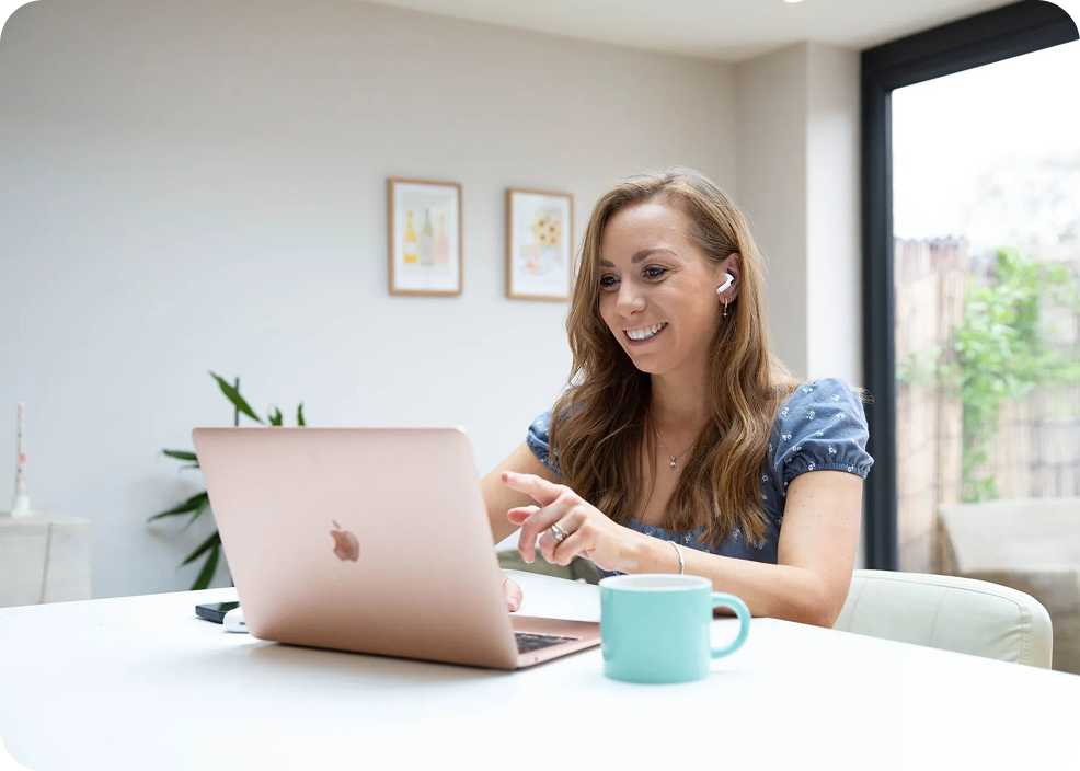 Woman smiling, working on a rose gold laptop at a white table, holding a coffee mug, indoors.