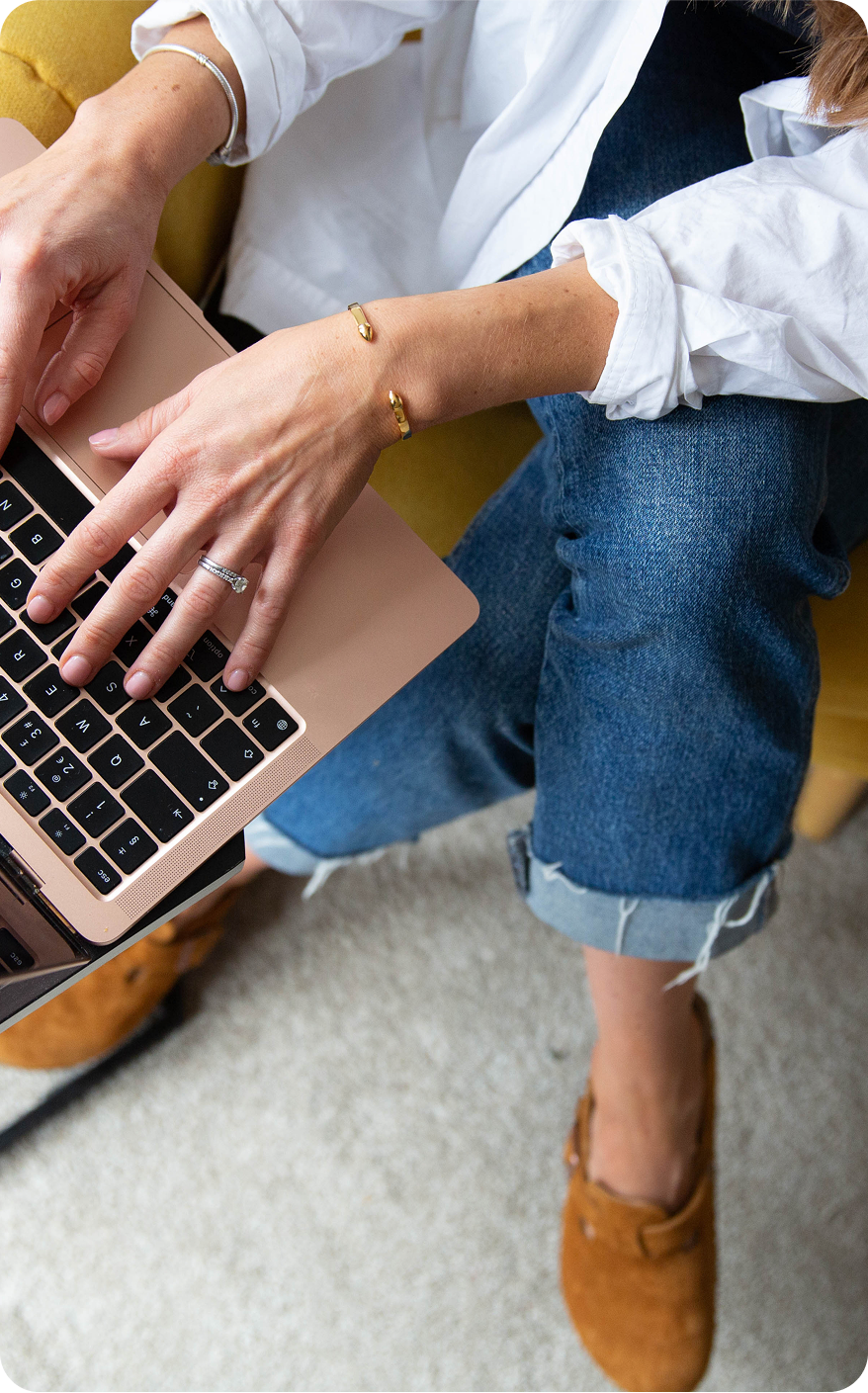 Person typing on a laptop, wearing a white shirt and jeans, sitting in a yellow chair.