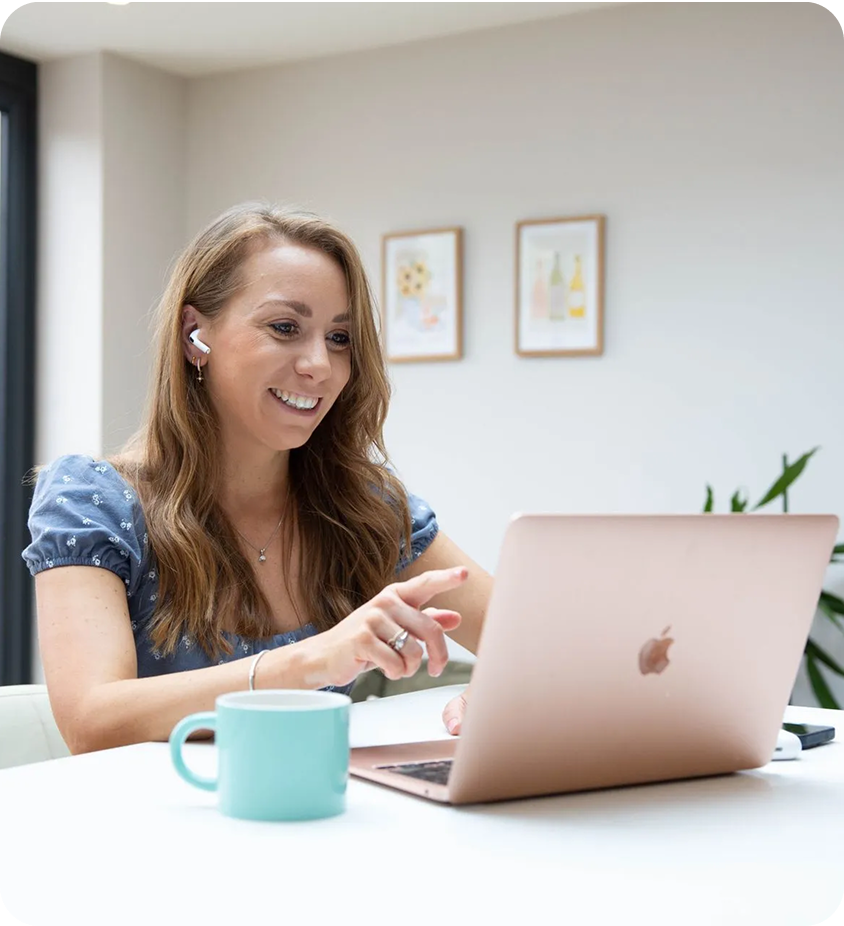 Woman with long hair smiles, working on a laptop at a white table. Wearing earbuds, blue dress, teal mug.