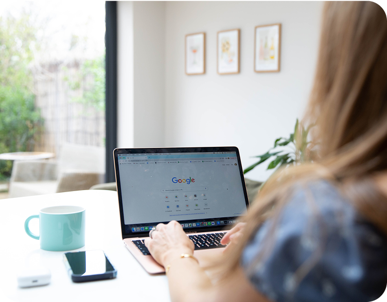 Woman using a laptop with Google search open, sitting at a white table. A mug and phone are next to the laptop.