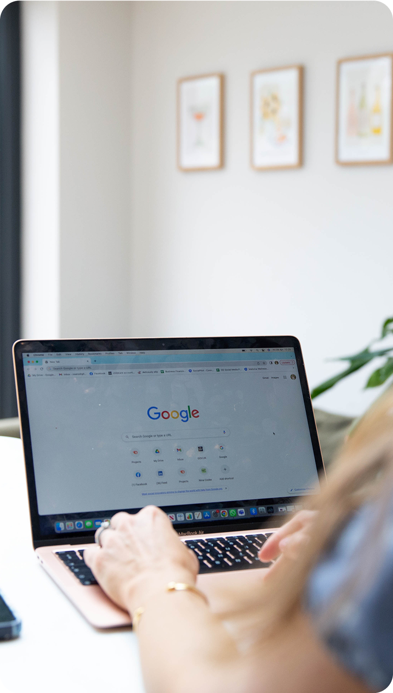 Person using a laptop with Google search open, working at a desk in a bright room with artwork on the wall.