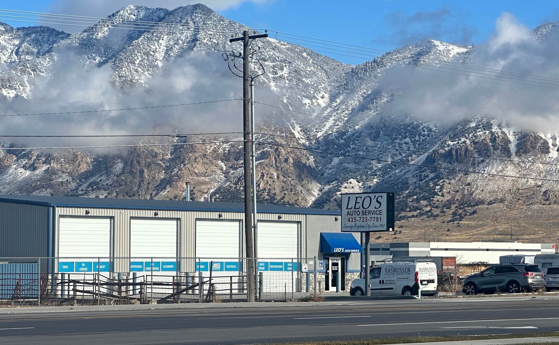 Snowy mountains behind a small industrial yard with trucks and blue-roofed buildings in winter. | Leo's Auto Service