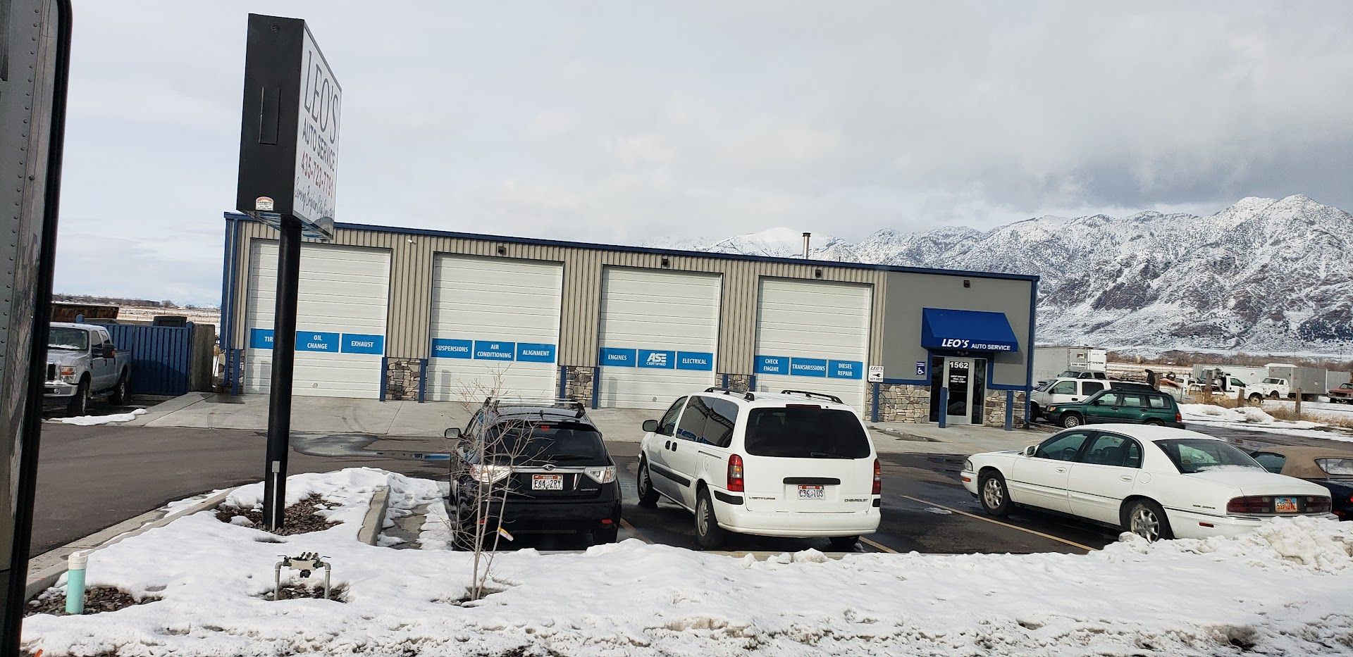 Snow-covered parking lot with three cars in front of a building, mountains in the background.| Leo's Auto Service