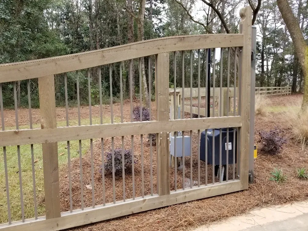 Tan wooden gate with vertical slats blocking a fenced yard with trees and dry grass beyond