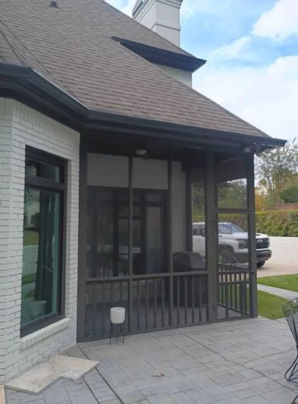 Covered front porch with dark railings and a gray shingled roof beside a house and driveway.