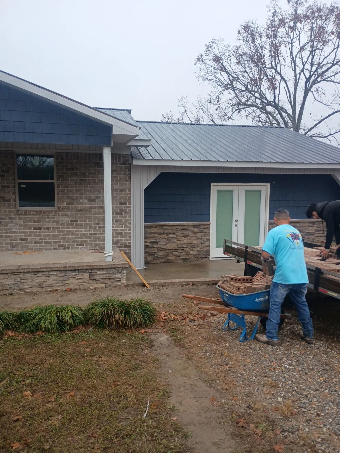 Workers near a brick house loading materials from a blue trailer on a gravel driveway
