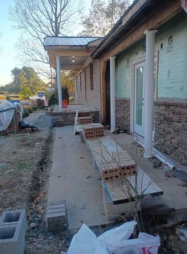 Brick house porch under construction with stacked bricks, boards, and a concrete walkway.