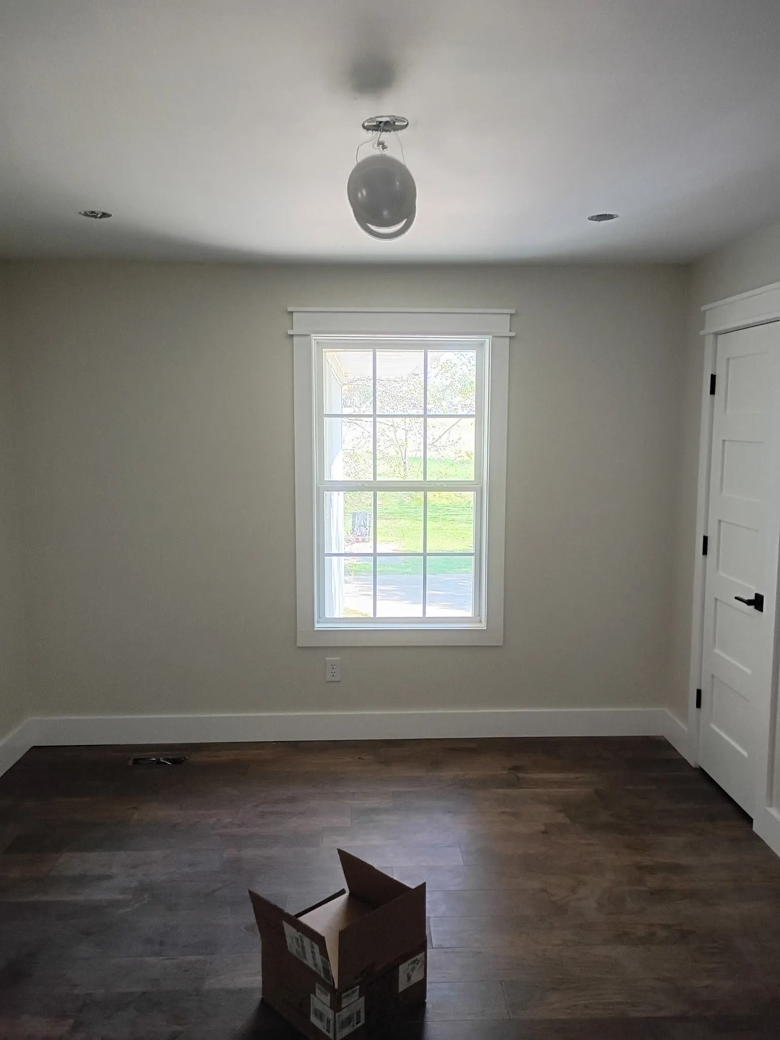 Empty white room with a window, dark wood floor, and an open cardboard box in the center.