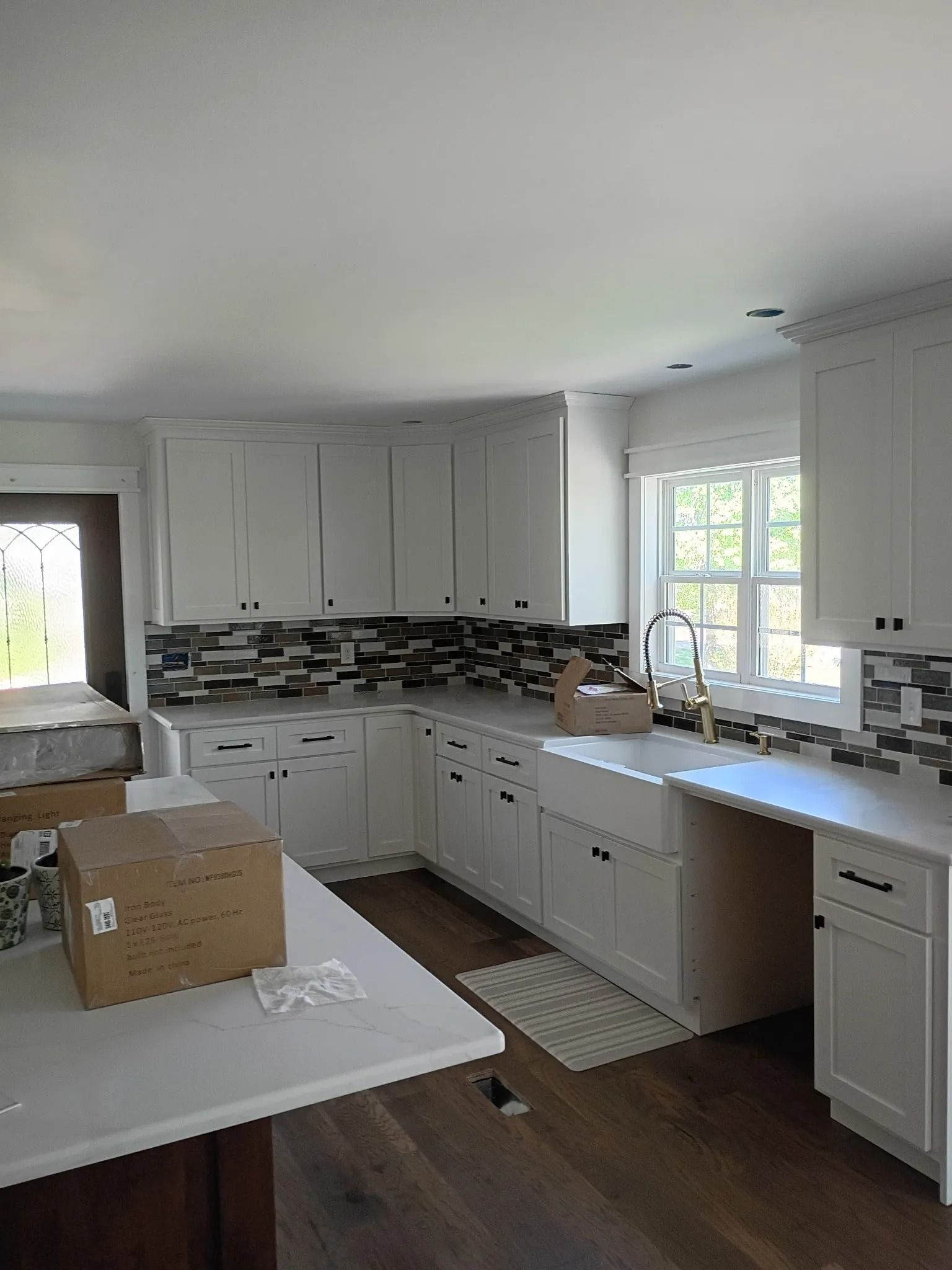 Bright kitchen with white cabinets, dark mosaic backsplash, and a box on the island in front of a window