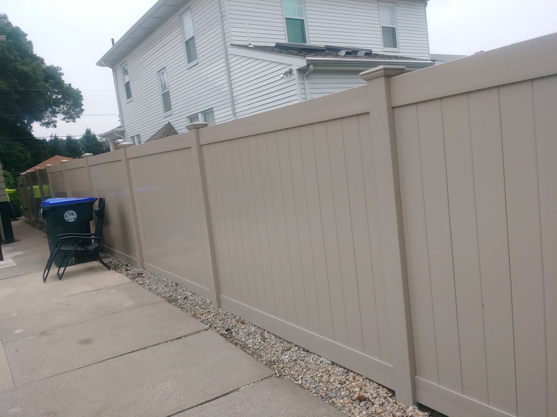 Tan vinyl fence next to a gray concrete sidewalk and a building. A blue trash bin sits near the fence.