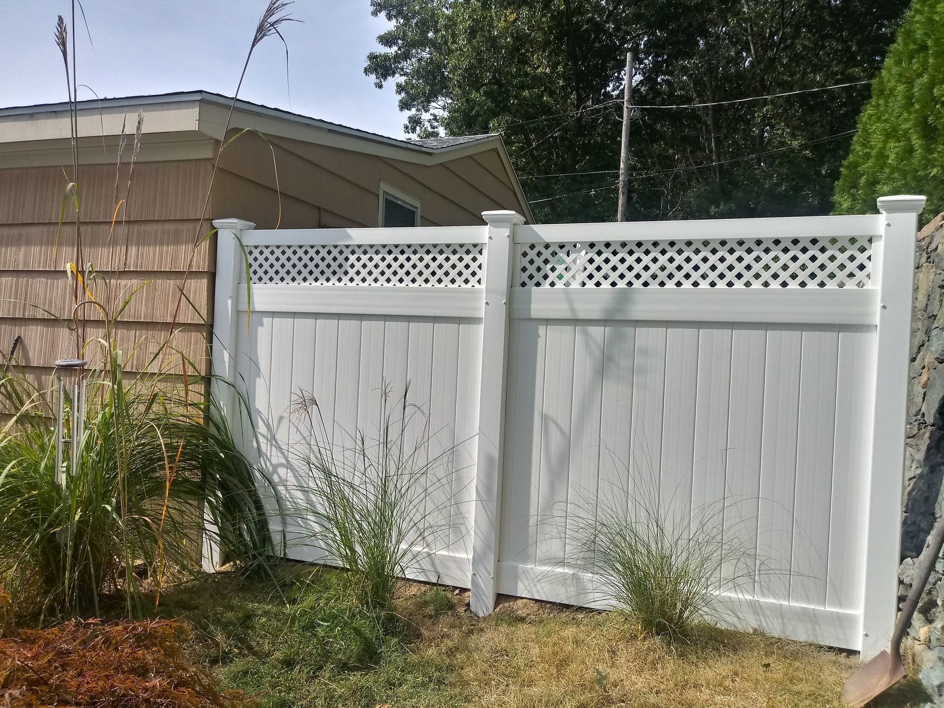 White vinyl fence with lattice top, in a grassy yard, next to a beige building with a dark roof.