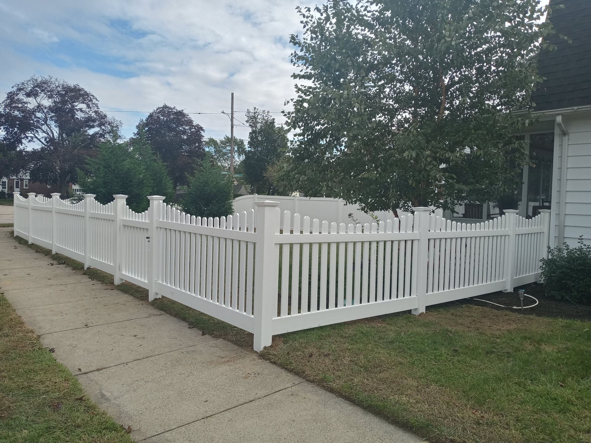 White picket fence bordering a sidewalk and lawn, beside a house with a cloudy sky.